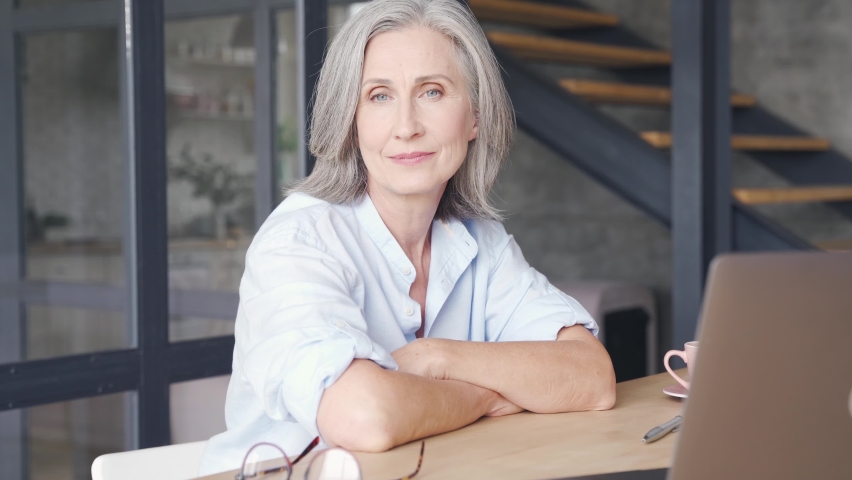 Smiling mature grey-haired business woman sits at workplace table. Confident middle aged lady, senior female professional coach, older executive leader close up face headshot portrait in office. Royalty-Free Stock Footage #1062406906