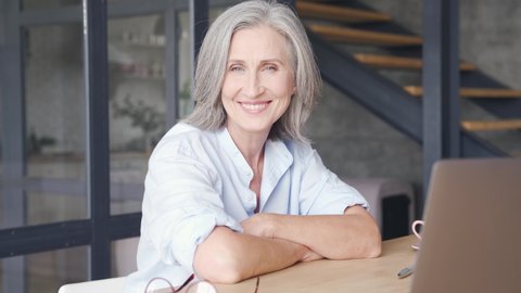 Smiling mature grey-haired business woman sits at workplace table. Confident middle aged lady, senior female professional coach, older executive leader close up face headshot portrait in office.