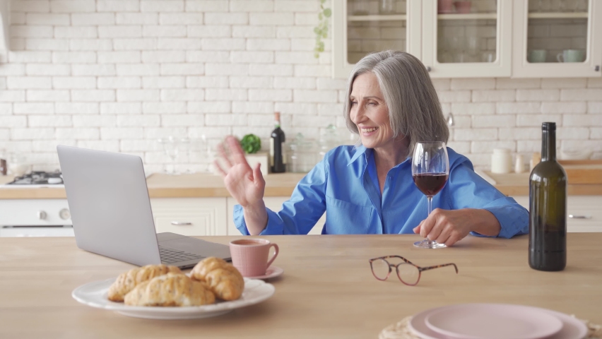 Happy senior lady drinking wine video calling friend on laptop at home. Smiling older woman holding glass saying toast talking by online social distance chat holiday party sitting at kitchen table. Royalty-Free Stock Footage #1062408172