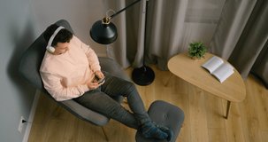 Young Man Talks Online with Woman by Video Call on Smartphone at Home at Night. Top View of Male Person in Headphones Sitting in Armchair in Cozy Room. COVID-19 Self-isolation Concept. 4K Static Shot