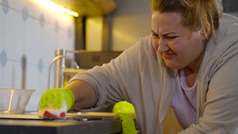 Close up of irritated young woman trying to wipe dirty spot on countertop with sponge. Overweight housewife on rubber gloves cleaning kitchen after cooking dinner