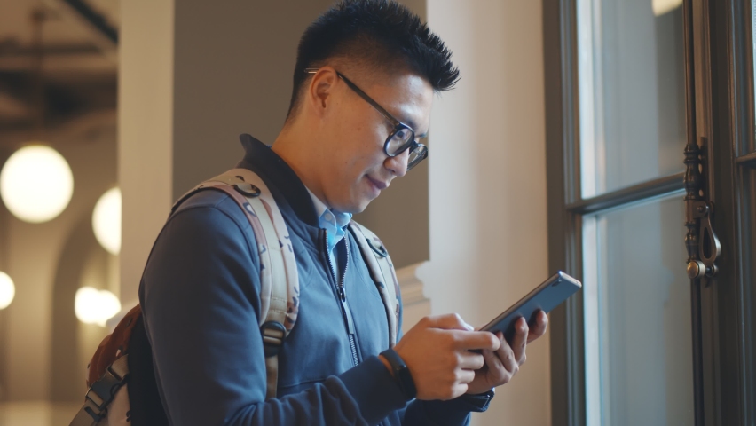 Smiling male college student checking mobile phone in campus building. Side view of cheerful asian college guy surfing internet or chatting on smartphone standing near window in classroom Royalty-Free Stock Footage #1062412624
