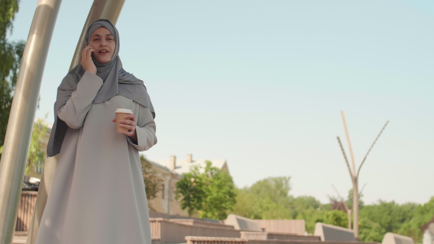 Medium shot of young Muslim woman wearing grey hijab is standing outdoors with coffee paper cup in her hands and chatting on telephone