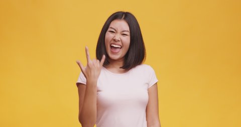 Carefree delighted asian woman demonstrating rock and roll hand gesture, punk sign, orange studio background