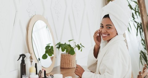 Portrait of happy attractive african american woman applying cream on face and smiling at camera, sitting after shower in bathroom, slow motion