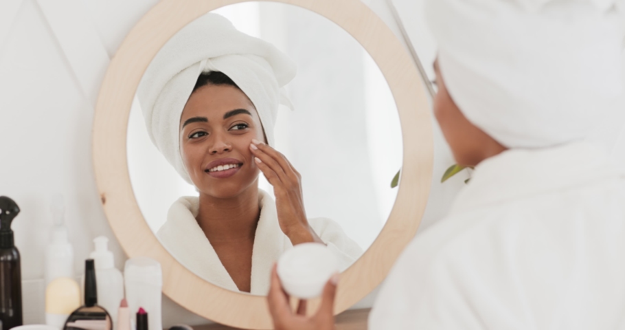Feminine daily procedures. Young african american lady in bathrobe applying cream on face, moisturizing skin after morning shower, slow motion | Shutterstock HD Video #1062422659