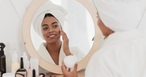 Feminine daily procedures. Young african american lady in bathrobe applying cream on face, moisturizing skin after morning shower, slow motion