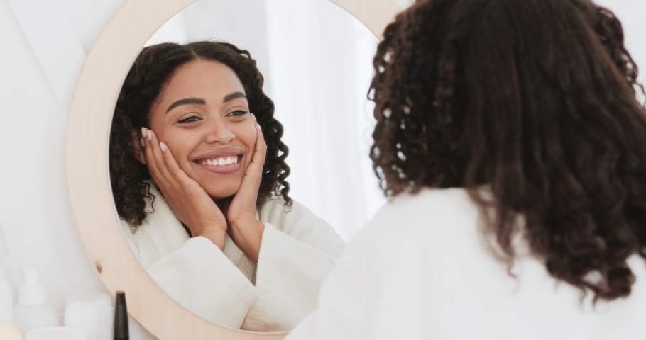 Love yourself. Happy beautiful black lady looking at mirror, smiling to her reflection, enjoying her appearance and home beauty procedures, slow motion