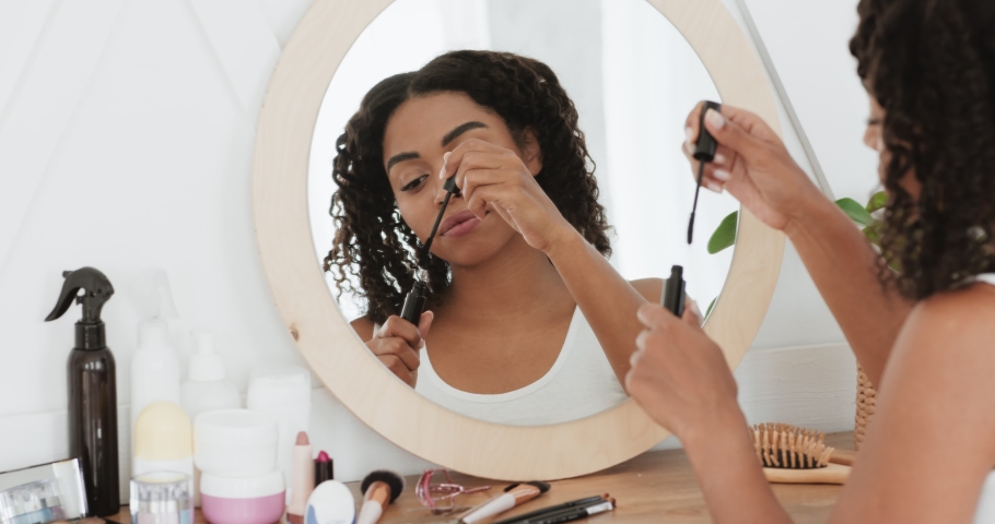 Daily make up. Young attractive black lady applying mascara on her eyelashes, looking at mirror at home, slow motion Royalty-Free Stock Footage #1062422686