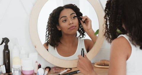 Daily make up. Young attractive black lady applying mascara on her eyelashes, looking at mirror at home, slow motion