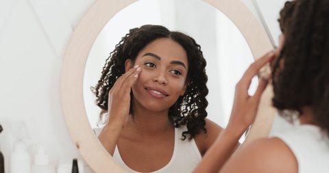 Young black healthy lady looking at mirror at home, touching her face and enjoying her skin condition, slow motion. Beauty and skincare concept