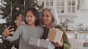 Joyous senior grandma and cheerful little girl of Asian ethnicity holding gift boxes, smiling and waving at smartphone camera while taking selfie or video calling beside Christmas tree at home