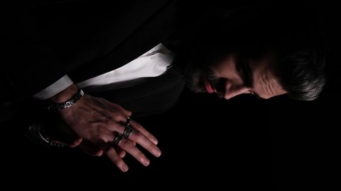 sexy elegant man touching fingers, looking to side and thinking, arranging black tuxedo, sensually grabbing neck and posing in a fashion light on dark background in studio