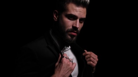 serious young man in black tuxedo crossing arms, holding hands in pockets, sensually looking to side and disappearing in the dark on black background in studio