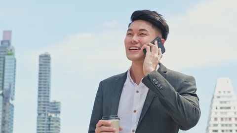 Asian young smart businessman talking in telephone in city. The man holding coffee cup walking outdoor to working office with smile face. Telecommunication and mobile network for business concept.