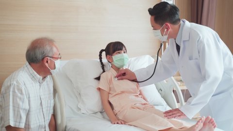 Asian doctor measure heart rate by stethoscope on little kid patient on bed in recovery room. Man and woman wearing mask having examination on girl during covid pandemic. Father taking care of child.