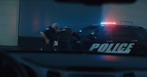 WIDE Police officer searches and handcuffs a suspect, African-American Black criminal. Police car lights flashing in the background. Shot with RED cinema camera and 2x Anamorphic lens