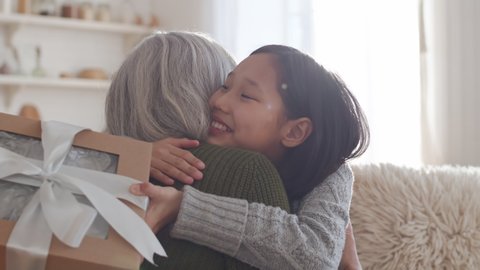 Happy Asian girl receiving Christmas gift from loving grandmother, getting surprised and giving hugs to her while sitting on sofa in living room