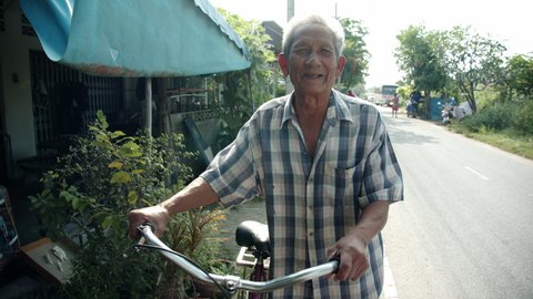 Slow motion scene of white-haired poor Asian elderly man , older than seventy stands smiling and is happily leading the bicycle on the roadside of his village