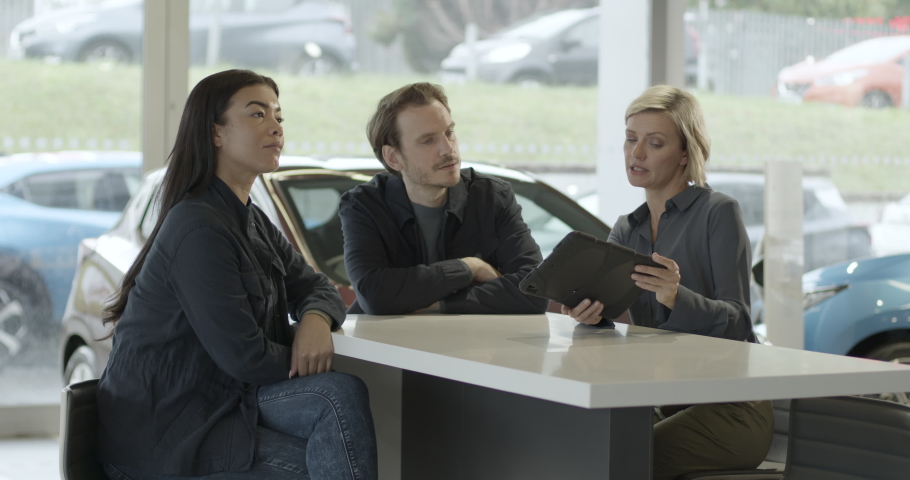 Saleswoman advising couple on car purchase in vehicle dealership showroom using digital tablet