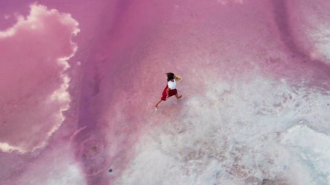 AERIAL, drone follows: happy laughing girl in red skirt runs on shallow water along white salty shore of amazing pink lake with clouds reflections. carefree female showing joy and happiness. – Stockvideo