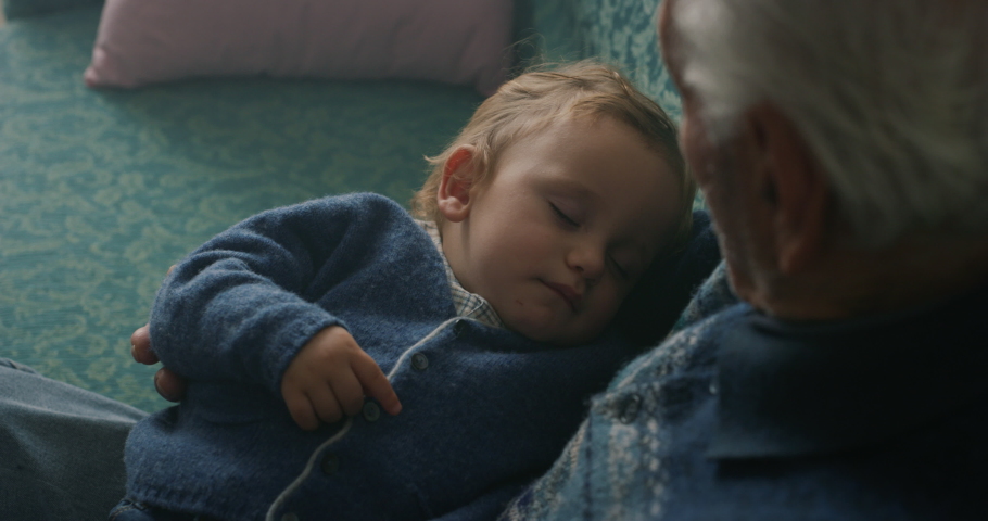 Cinematic close up shot of senior gray hair grandfather is cuddling grandson baby sleeping peacefully on arms while sitting on sofa at home.Concept:life, grandparents,love,care, generation, childhood Royalty-Free Stock Footage #1062429145