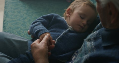 Cinematic close up shot of senior gray hair grandfather is cuddling grandson baby sleeping peacefully on arms while sitting on sofa at home.Concept:life, grandparents,love,care, generation, childhood