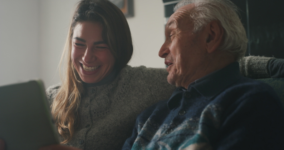 Cinematic shot of happy granddaughter and grandfather are having fun to make a selfie or video technology call to family and relatives with tablet while sitting on sofa in living room at home. Royalty-Free Stock Footage #1062429829