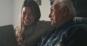 Cinematic shot of happy granddaughter and grandfather are having fun to make a selfie or video technology call to family and relatives with tablet while sitting on sofa in living room at home.