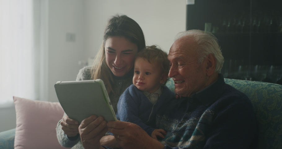 Cinematic shot of happy family: grandfather, daughter and grandson baby having fun to make selfie or video technology call to family and relatives with tablet or smartphone on sofa in living room. Royalty-Free Stock Footage #1062430066