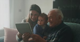 Cinematic shot of happy family: grandfather, daughter and grandson baby having fun to make selfie or video technology call to family and relatives with tablet or smartphone on sofa in living room.
