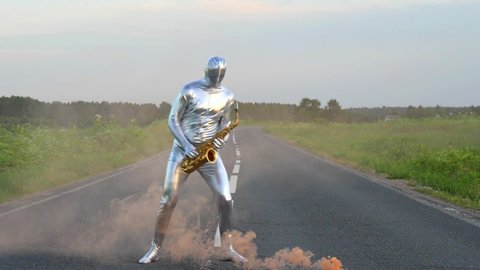 Man musician saxophonist in full body-hugging silver and electric suit holding golden alto saxophone, standing and dancing on empty road in summer with perspective. Freak, alien, UFO, Orange smoke.