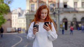 Attractive young woman with red hair hold coffee use phone smiling. Walking on street in city center. Travel tourist cellphone outdoors. Portrait. Slow motion - Powered by Shutterstock - Get 15% off with code: PIKWIZARD15
