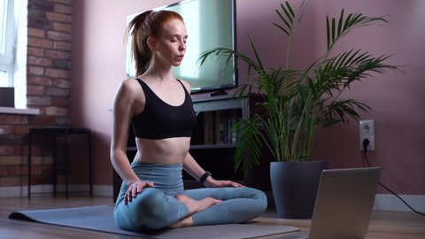 Calm woman sitting in lotus pose with closed eyes near laptop at home meditating alone on floor. Female performs deep relaxing breathing exercises at living room. Lady making deep breath-exhalation. - Βίντεο στοκ