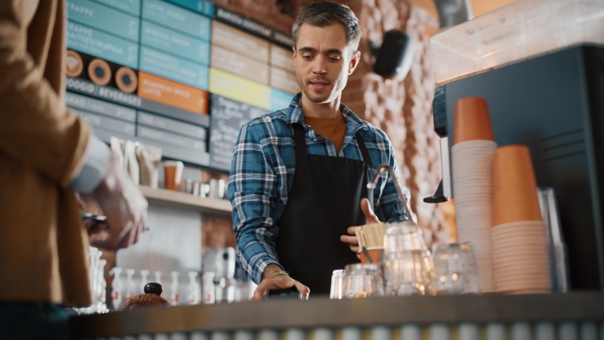 Tall Caucasian Customer Pays for Coffee and Pastry with Contactless NFC Payment Technology on Smartphone to a Handsome Barista in Blue Checkered Shirt. Contactless Mobile Payment in Cafe Concept. Royalty-Free Stock Footage #1062436996
