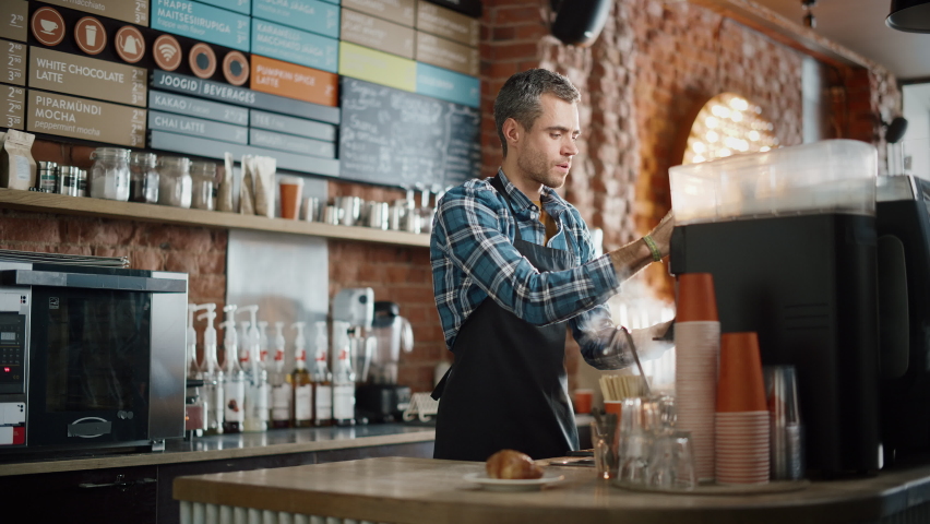 Handsome Barista in Checkered Shirt and Black Apron is Making a Cup of Tasty Cappuccino in Coffee Shop Bar. Portrait of Happy Employee Behind Cozy Loft-Style Cafe Counter. Royalty-Free Stock Footage #1062437020