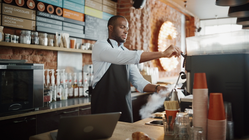Handsome Black Afro-American Barista Wearing Apron is Making a Cup of Tasty Cappuccino in Coffee Shop Restaurant. Portrait of Happy Employee Behind Cozy Loft-Style Cafe Counter. Royalty-Free Stock Footage #1062437032