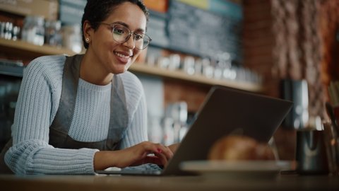 Young and Beautiful Latina Small Business Owner is Working on Laptop Computer and Checking Inventory in a Cozy Cafe. Happy Restaurant Manager or Employee Browsing Internet and Chatting with Friends.