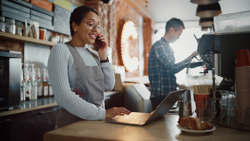 Latin American Coffee Shop Employee Accepts a Pre-Order on a Mobile Phone Call and Writes it Down on Laptop Computer in a Cozy Cafe. Restaurant Manager Browsing Internet and Talking on Smartphone. Royalty-Free Stock Footage #1062437053