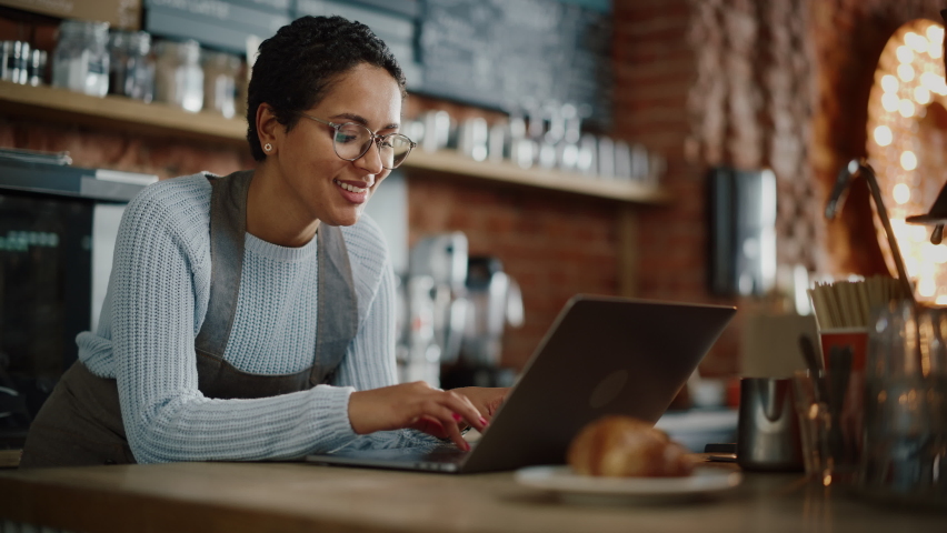 Young and Beautiful Latina Coffee Shop Owner is Working on Laptop Computer and Checking Inventory in a Cozy Cafe. Happy Small Business Owner or Employee Browsing Internet and Chatting with Friends. Royalty-Free Stock Footage #1062437056