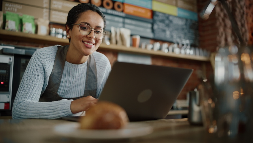 Young and Beautiful Latina Coffee Shop Owner is Working on Laptop Computer and Checking Inventory in a Cozy Cafe. Happy Restaurant Manager or Employee Browsing Internet and Chatting with Friends. Royalty-Free Stock Footage #1062437059
