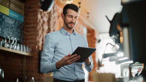 Handsome Caucasian Small Business Owner is Working on Tablet Computer and Checking Inventory in a Cozy Loft-Style Cafe. Successful Restaurant Manager Browsing Internet Behind a Barista Counter.