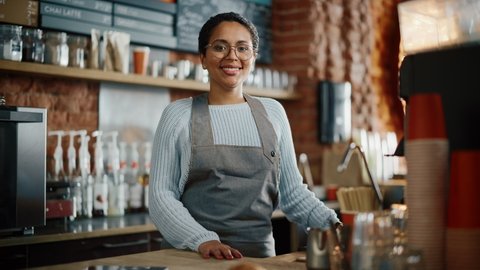 Beautiful Latin American Female Barista with Short Hair and Glasses is Projecting a Happy Smile in Coffee Shop Bar. Portrait of Happy Employee Behind Cozy Loft-Style Cafe Counter in Restaurant.