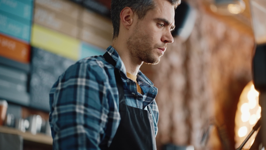 Close Up Portrait of a Handsome Caucasian Male Barista in Checkered Shirt Making Cappuccino in a Coffee Shop Bar. He Pours Fresh Coffee from a Machine and Warms Up the Milk at a Cozy Loft-Style Cafe. Royalty-Free Stock Footage #1062437128