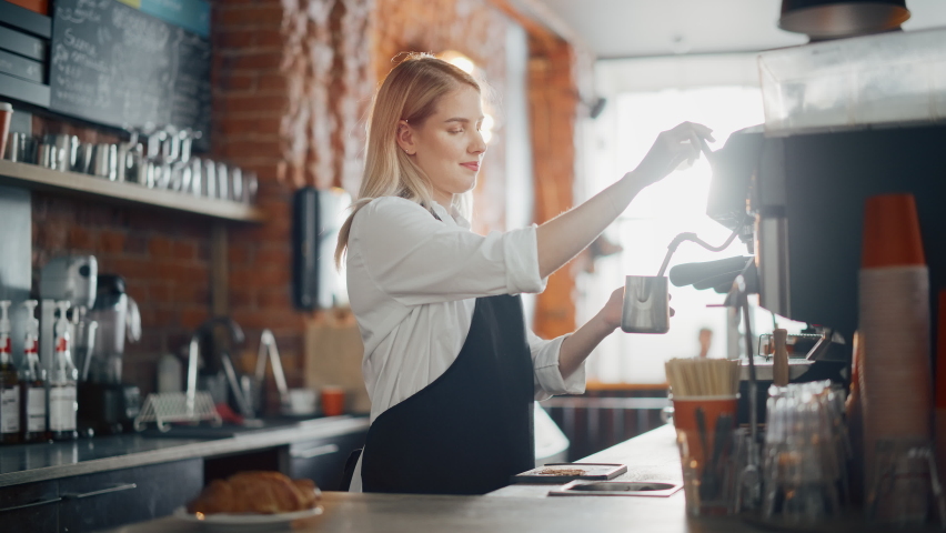 Beautiful Young Caucasian Barista with Blond Hair is Making a Cup of Fresh Coffee in a Cafe. Happy and Smiling Bar Employee Posing while Working in a Cozy Loft-Style Coffee Shop Restaurant Counter. Royalty-Free Stock Footage #1062437143