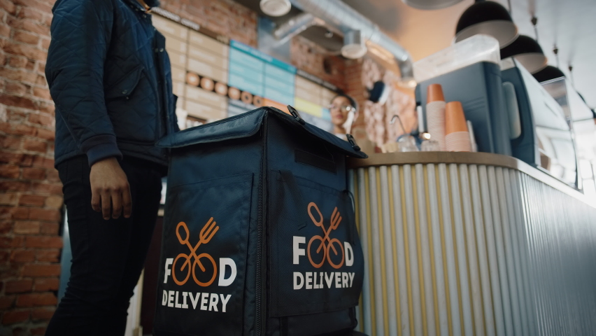 Beautiful Happy Latin Barista Serves Order to a Food Delivery Courier Picking Up Two Take Away Coffees and Pastries from a Cafe Restaurant. Delivery Guy Puts Food in His Hot Thermal Insulated Bag. Royalty-Free Stock Footage #1062437188