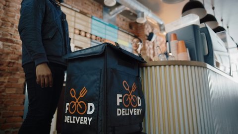 Beautiful Happy Latin Barista Serves Order to a Food Delivery Courier Picking Up Two Take Away Coffees and Pastries from a Cafe Restaurant. Delivery Guy Puts Food in His Hot Thermal Insulated Bag.