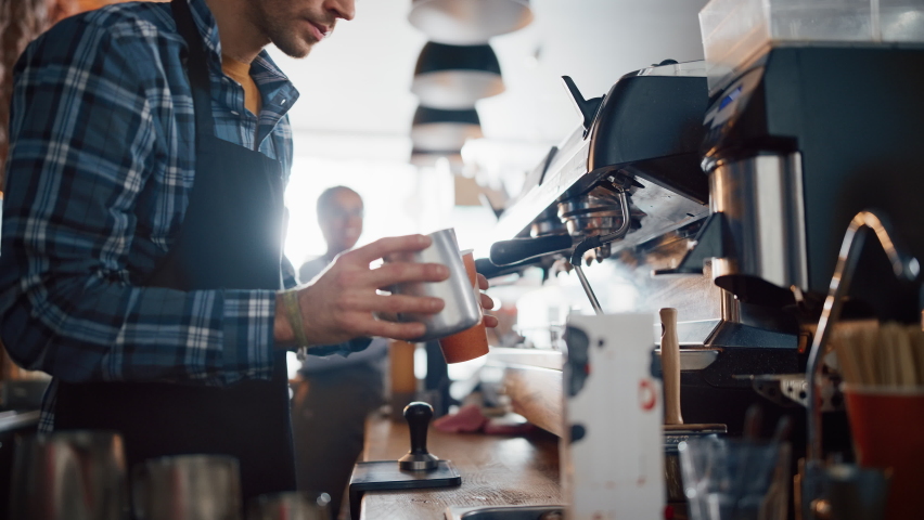 Handsome Male Barista in Checkered Shirt is Making a Take Away Cappuccino in Disposable Recycle Cup for a Customer in a Coffee Shop Bar. Female Cashier Works at a Cozy Cafe Counter in the Background. Royalty-Free Stock Footage #1062437206
