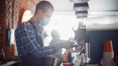 Male Barista in Face Mask is Making a Cappuccino in a Coffee Shop Bar. Lockdown Social Restrictions Concept During Covid-19 Pandemic in Restaurants. Female Cashier Works at a Cafe in the Background.