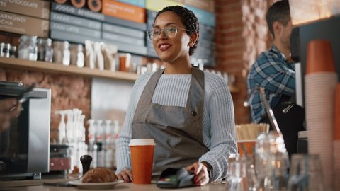 Joyful Multiethnic Diverse Woman Gives a Payment Terminal to Customer Using NFC Technology on Smartphone. Customer Uses Mobile to Pay for Take Away Latte and Pastry to a Barista in Coffee Shop.